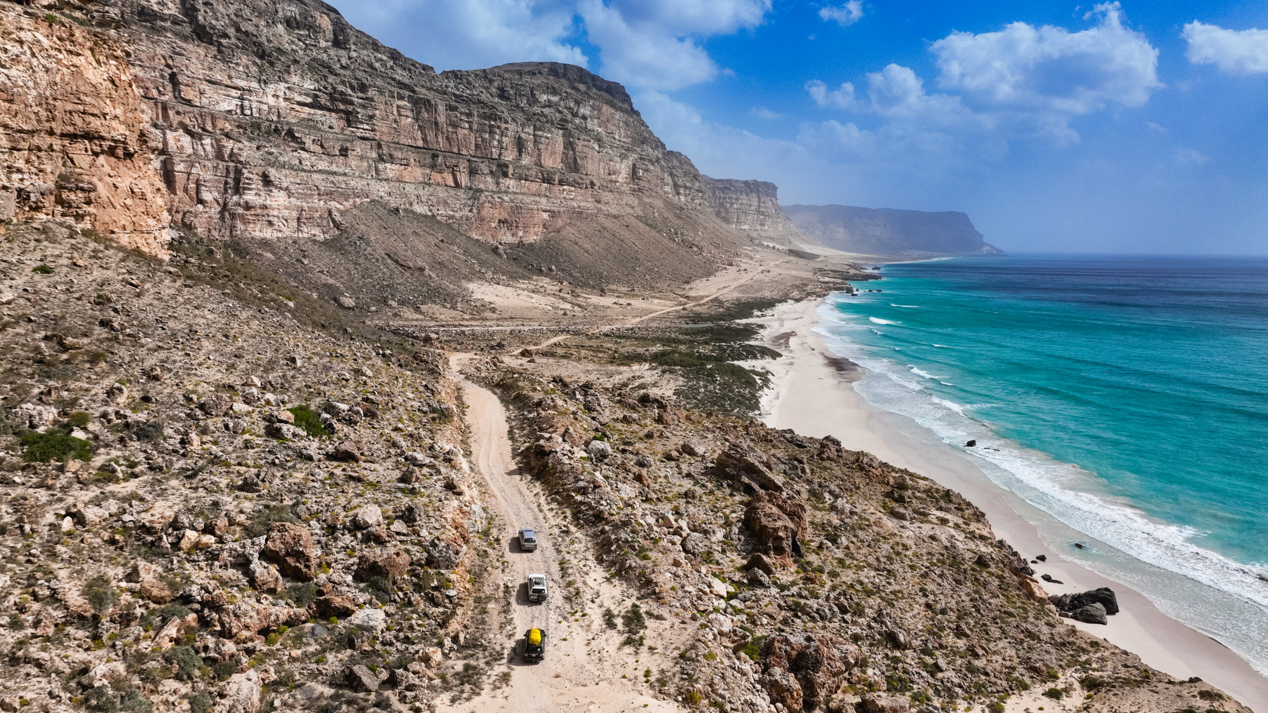 Off road vehicles navigate sandy paths along the stunning coastline of Socotra Island, showcasing unique geological formations.